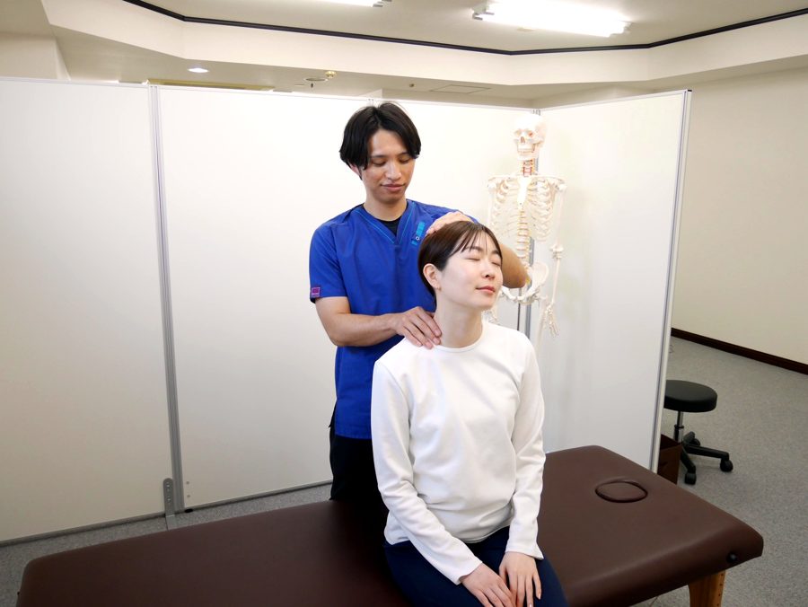 座位の患者さんの首をやさしく施術する小林院長（骨格模型のある施術室）